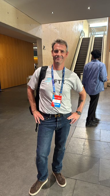 A smiling man in a gray FTK t-shirt and conference lanyard stands inside what appears to be the Ethereum Blockchain Convention venue, with escalators visible behind him, perfectly capturing the excitement of attending @EBlockchainCon.