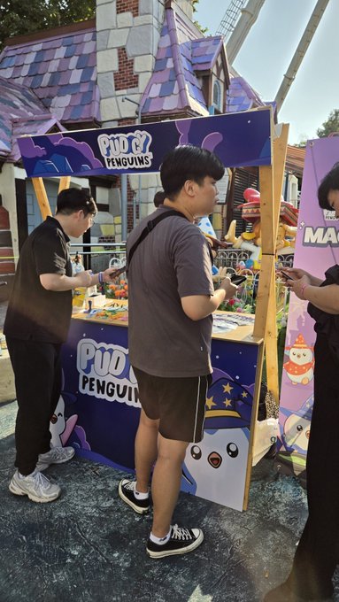 Three men stand at an outdoor booth decorated with purple graphics and 'PUO PENGUINS' branding, with a whimsical purple house structure in the background.
