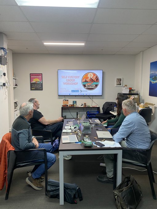 A group of people sit around a conference table in a meeting room watching a presentation displayed on a TV screen that reads 'SELF CUSTODY GROUP WORKSHOP' with 'BITCOIN FOR BEGINNERS JOURNEY' visible on the side.