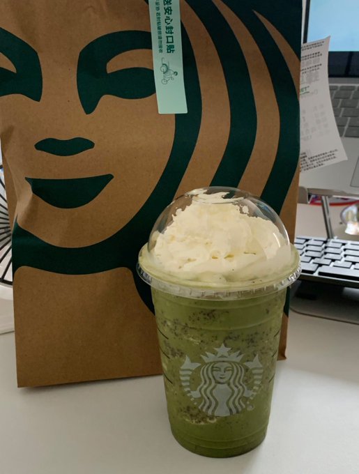 A Starbucks matcha Frappuccino topped with whipped cream sits on a desk next to a paper bag with the green Starbucks mermaid design and Korean text '주문&픽업은' (Order & Pickup).