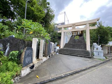 A photograph of a Japanese Shinto shrine entrance, featuring a white torii gate at the top of a stone staircase, flanked by stone monuments with inscriptions and surrounded by lush greenery.