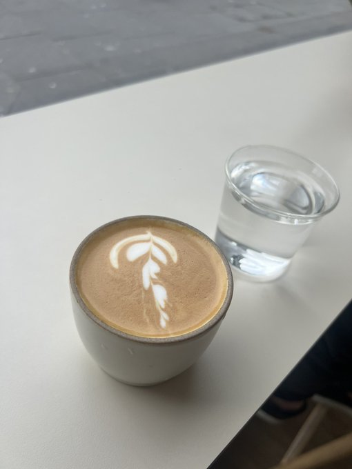 A cappuccino with leaf-patterned latte art in a ceramic cup sits on a white counter next to a glass of water.
