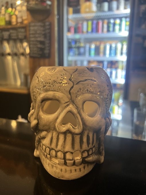A tan ceramic skull-shaped mug with hollow eyes and a crack down the forehead sits on a dark bar counter, with beverage taps and a lit refrigerator blurred behind it.