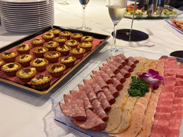 A buffet display featuring a tray of mini quiches or egg tarts alongside a platter of sliced cured meats (likely salami and ham) garnished with parsley and a purple orchid.