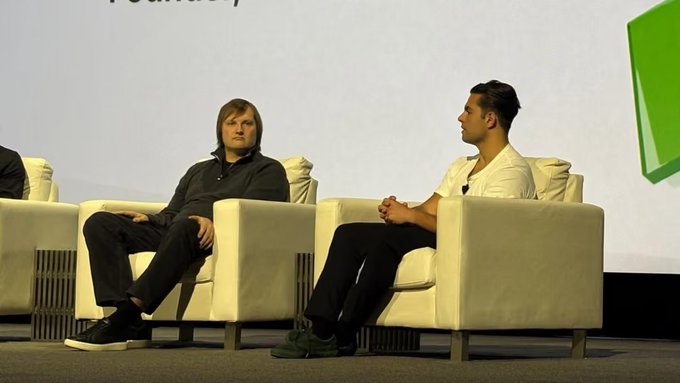 Two men sit in white armchairs on a stage, engaged in conversation during what appears to be a tech or business conference panel.