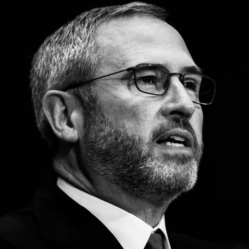 The image features a man wearing glasses and a suit jacket with a tie. He appears to be looking off into the distance, possibly deep in thought or contemplation. The background of the image is black, which contrasts with the white collar on his shirt. There are no other objects or people visible in the photo.
