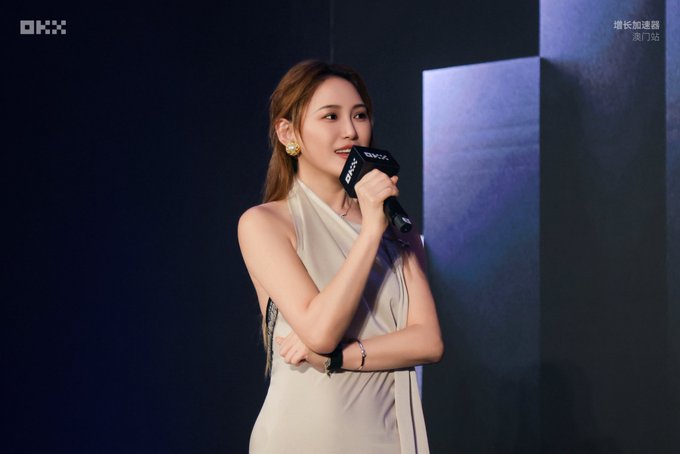 A young woman in a cream halter dress speaks into a microphone branded with "OKX" at what appears to be an OKX event in Macau (澳门站). The post mentions @dearcindyt among others, suggesting she is one of the professional hosts being praised for her elegance at the event.