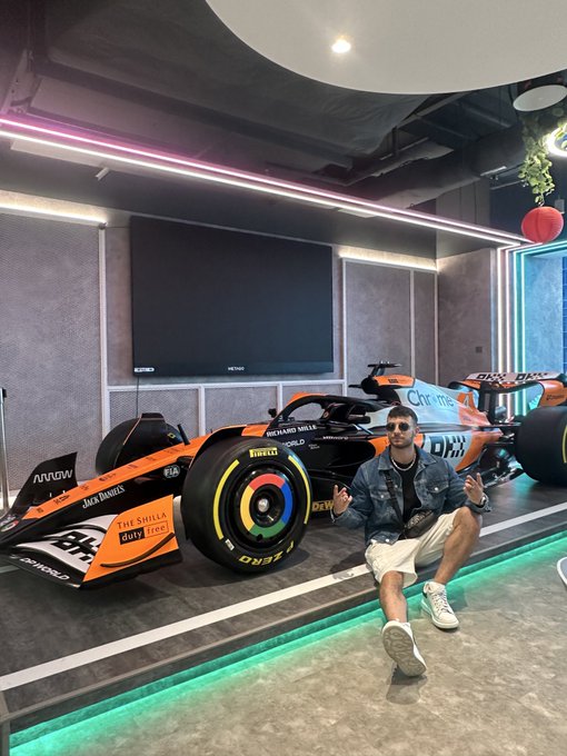A man in sunglasses and a denim jacket crouches excitedly next to a McLaren Formula 1 race car featuring "OKX," "Chrome," "Dell," and "Jack Daniel's" sponsorship branding, posing for a photo in what appears to be a stylish racing-themed lounge or showroom.