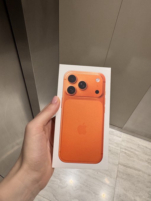 A hand holds a white box containing a new orange smartphone, likely an iPhone, displaying its three-camera system.