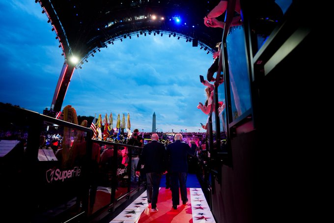 The image shows two men walking down a red carpet, with a crowd of people behind them. They are wearing suits and appear to be attending an event or ceremony. The crowd consists of at least thirteen individuals, some standing closer together while others have more space between them. The scene suggests that the man in front is likely the speaker or performer being introduced by the post.