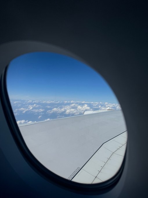 A view through an airplane window showing the aircraft's wing extending over a vast expanse of white clouds against a clear blue sky.