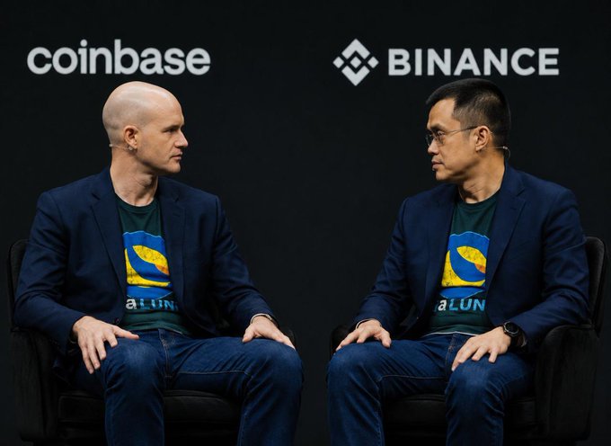The image shows two men sitting next to each other, both wearing blue suits with a logo on their shirts. They are positioned against a black background that features logos for Coinbase and Binance. The man on the left has his hands folded in front of him, while the man on the right is facing away from the camera. Both men appear to be engaged in conversation or observing something happening around them.