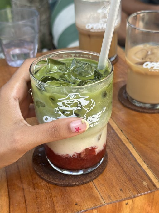 A hand with star-decorated nails holds an iced matcha drink in a glass featuring an 'OPEN HOUSE INDONESIA' logo, sitting on a wooden table alongside other beverages.