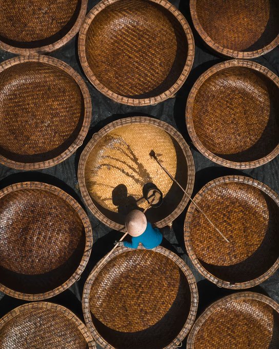 An aerial overhead photograph showing a worker wearing a conical hat weaving a large bamboo basket among many similar round woven baskets laid out in a pattern.