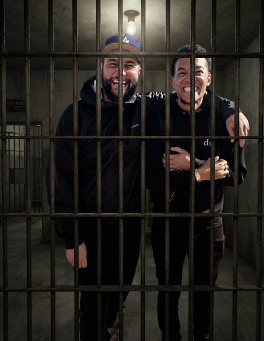 Two men smiling and laughing behind the bars of a prison cell set; one wears a black jacket and a blue LA Dodgers cap, the other a black shirt.