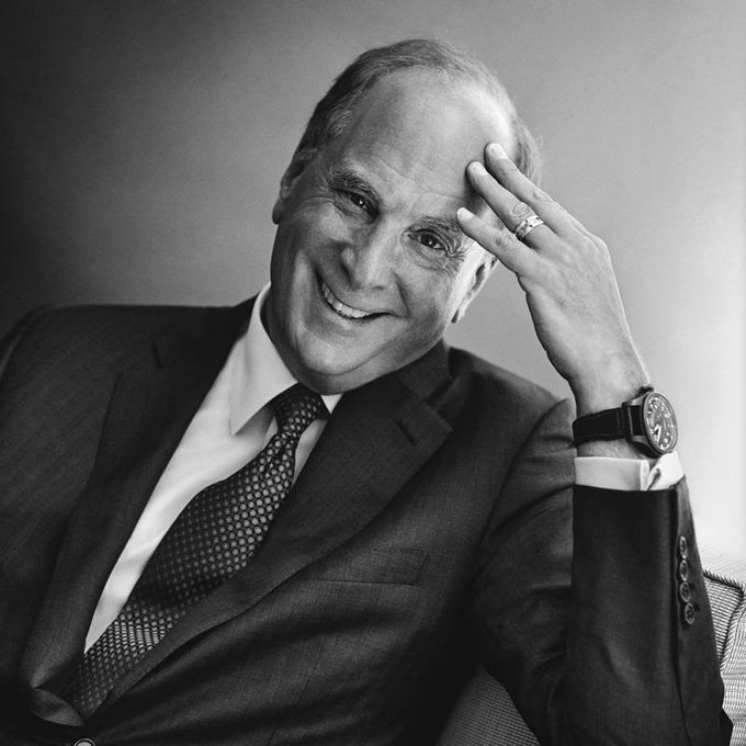 A black and white portrait of a smiling older man in a suit and tie, resting his hand on his forehead, wearing a watch and a ring.