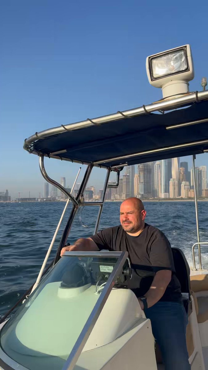 The image features a man sitting inside a boat with his hands on the steering wheel, looking out at the water. The boat appears to be a motorboat or yacht, as indicated by its design elements such as the windshield and the presence of a small cabin area. In the background, there are buildings that suggest an urban setting, possibly in Dubai, as suggested by the post. The man's position on the boat and his focus on the water imply that he is enjoying the experience of being out on the sea or lake.