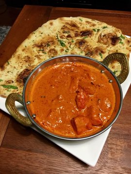 A metal karahi bowl filled with orange-red paneer butter masala curry sits on a white square plate beside pieces of naan flatbread, served on a wooden tray.