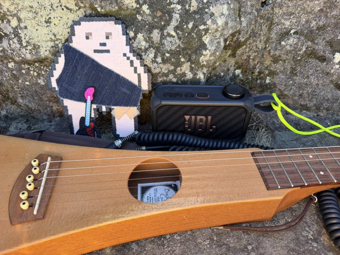 A pixel art figurine of a yeti or ape-like creature stands next to a black JBL portable speaker, with a wooden ukulele resting in the foreground against a rocky background.