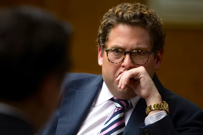 A man with curly brown hair, wearing tortoiseshell glasses, a navy suit, pink shirt, and striped tie, sits with his hand to his mouth in a thoughtful pose.