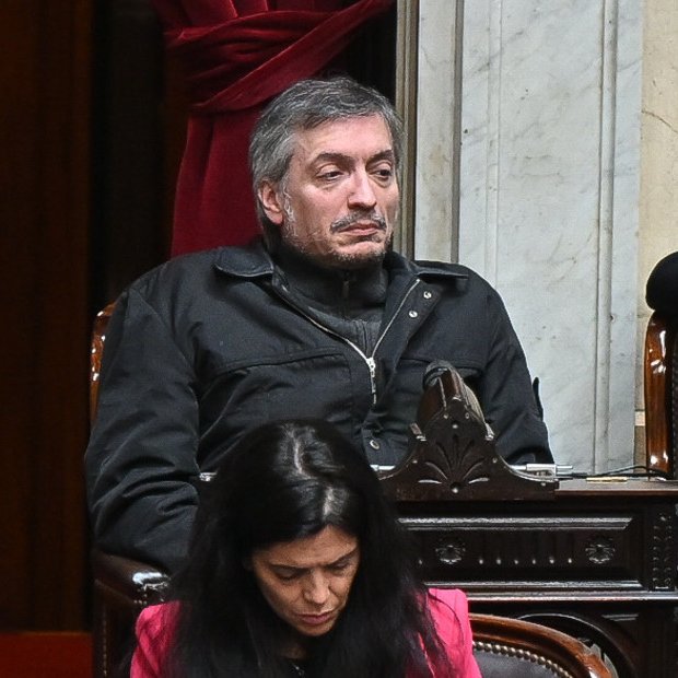 A man with a mustache in a dark jacket sits in an ornate wooden chair, looking to the side, with a woman in pink visible below him in a formal setting.