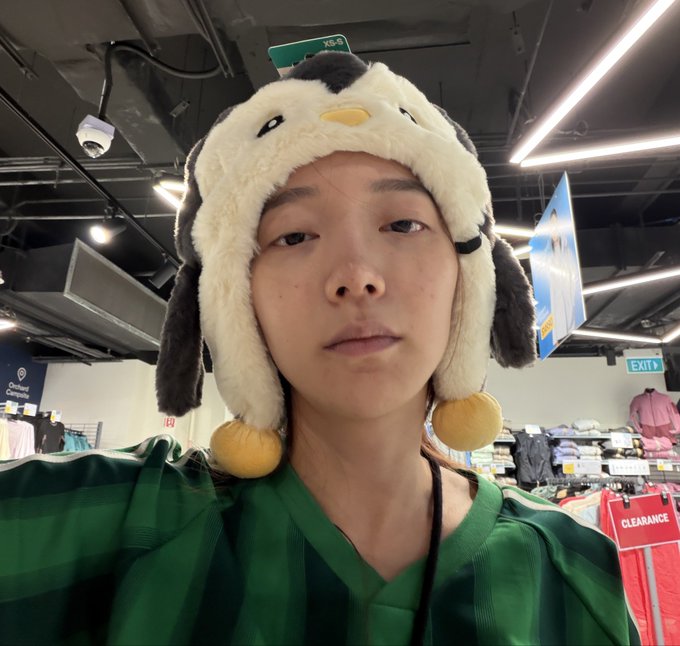A person wearing a plush penguin trapper hat poses inside a retail store, with "KB-6," "EXIT," and "CLEARANCE" signs visible in the background. The cozy indoor hat playfully reinforces the caption's joke about "degens" (likely crypto/enthusiast slang) not being suited for the outdoors.