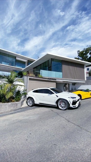 A modern two-story house with large glass windows and a balcony, with a white Lamborghini Urus SUV and a yellow sports car parked in the driveway.