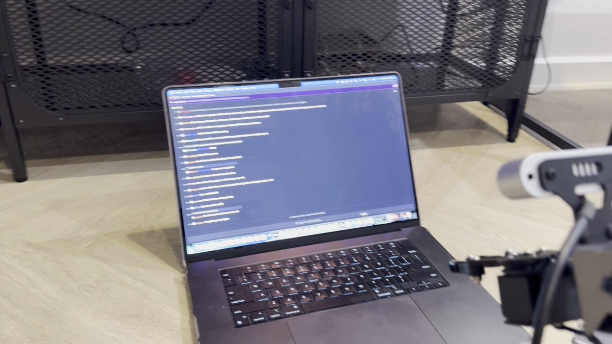 The image shows a laptop computer sitting on a wooden floor with its screen open displaying some text or code. The laptop appears to be turned off, possibly indicating that it's not currently being used for any tasks at the moment. The background of the image features a black cabinet and a white wall, providing a simple yet functional workspace.