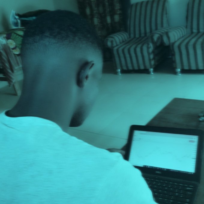 A young man in a light shirt sits on the floor working on a Dell laptop, with striped armchairs and curtains visible in the background.