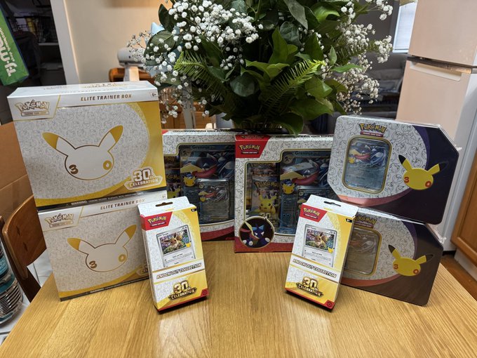 A collection of Pokémon TCG products displayed on a wooden table, including an Elite Trainer Box and booster packs featuring Pikachu and other Pokémon artwork, with a floral arrangement in the background.