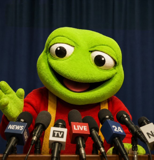 Kermit the Frog puppet character sitting at a podium with multiple microphones labeled 'NEWS', 'TV', 'LIVE', and '24A', appearing to give a press conference.