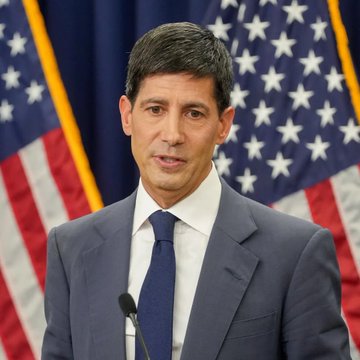 A man in a dark suit and blue tie speaking at a podium, standing in front of American flags.