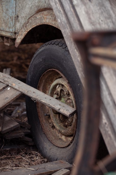 A close-up photograph of an old, rusted tire and wheel of a weathered vehicle, with a wooden plank resting across it and more wooden debris scattered around.
