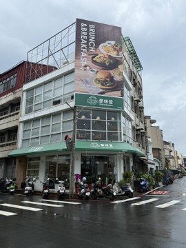 A street corner on a rainy day features a brunch café with a large rooftop sign reading "BRUNCH BREAKFAST" alongside food images, with scooter riders waiting at the crosswalk under a green awning marked "果核豆" — matching the post's lament about endless drizzly weather with no sun in sight.
