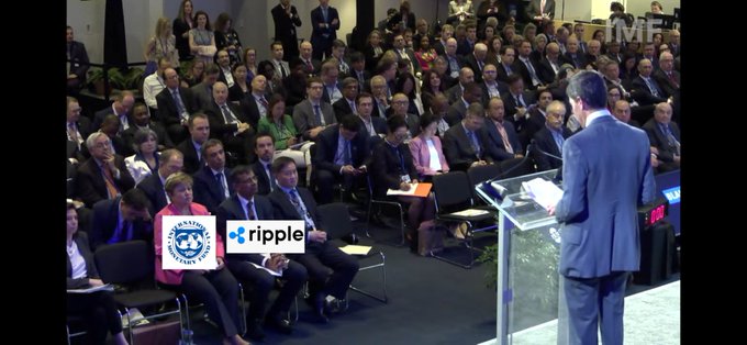 In the image, there are two men standing behind a podium, one of whom appears to be giving a speech at an event. The audience consists of numerous people sitting in rows of chairs, attentively listening to the speaker. A sign can be seen on the stage that reads "Ripple". Additionally, there is a text overlay in the image that says "The new Fed governor speaking at a irmf conference with Ripple & IMF right in the front seated next to each other." This suggests that the event might be related to cryptocurrency or financial technology.