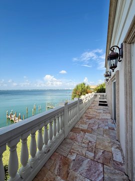 A scenic oceanfront view from a stone balcony with white balustrades, overlooking turquoise waters and a wooden dock under a blue sky.
