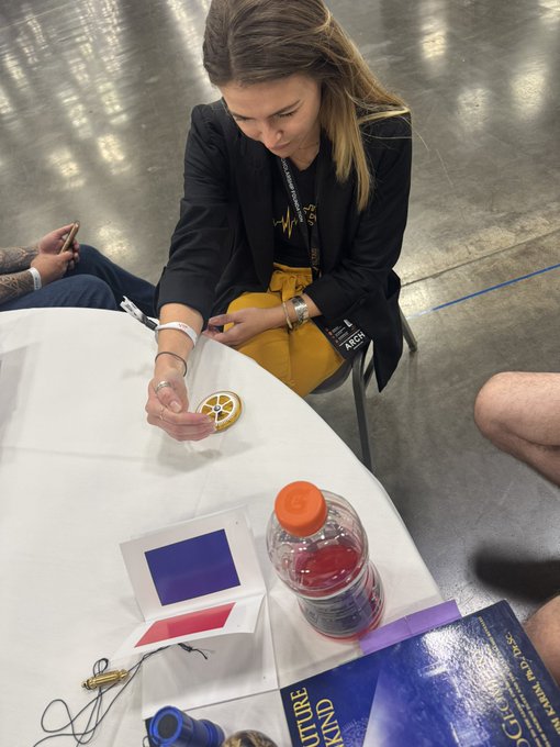 A young woman with long brown hair sits at a round white table in a large hall, focused on a craft project with red string, surrounded by bottles and patterned cards, possibly at a convention.