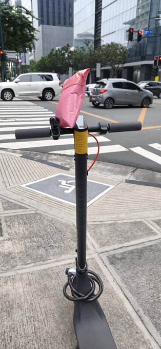 An electric scooter parked on a city sidewalk with a pink bubble-wrapped package resting on its handlebars, near a crosswalk and parked cars.