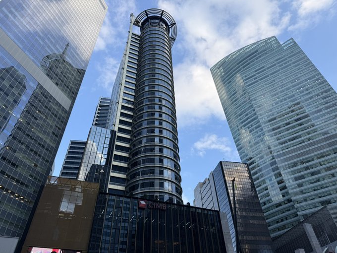 A low-angle view of modern skyscrapers against a blue sky, featuring a distinctive cylindrical tower and the 'CIMB' bank logo on a lower building.