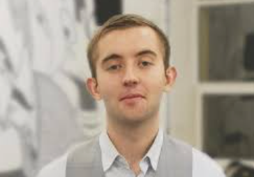 A young man with short light brown hair, wearing a gray vest over a white collared shirt, is captured mid-speech with a slight smile, likely delivering a Chainlink-related announcement tied to SmartCon events such as the CCIP or CRE technical developments referenced in the post.