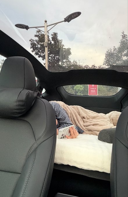 A person relaxes on a white mattress in the back of a car, wrapped in a beige blanket and holding a phone, with a panoramic sunroof open to the sky above. The view outside includes trees, street lamps, and a red sign reading "TULALIP" through the rear window, highlighting the roomy and sky-lit comfort the wife enjoys.