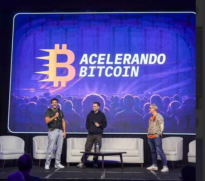 Three men stand on a stage in front of a large screen displaying a Bitcoin logo and the text 'ACELERANDO BITCOIN' in Portuguese, with an audience visible in the background.