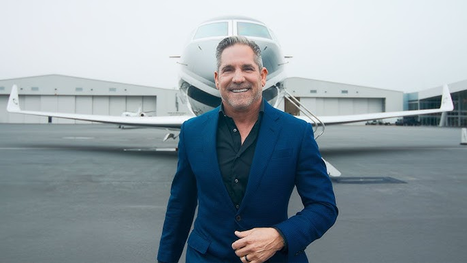A smiling man in a blue blazer stands in front of a private jet on a tarmac, posing confidently for the camera.