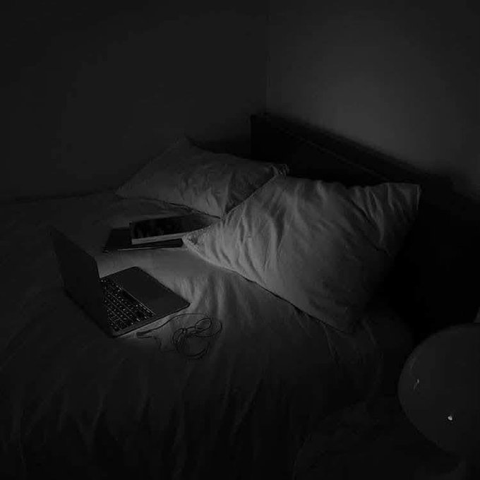 A dimly lit bedroom with a laptop and a phone on an unmade bed with white pillows.