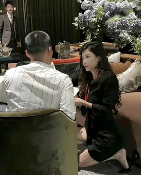 A woman with long dark hair wearing a black outfit sits on a couch facing a man in a striped shirt, appearing to be in a meeting or conversation in an indoor venue.