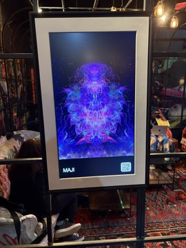 A digital art display labeled "M.A.I." showing a vibrant neon blue and purple abstract character on a screen mounted in what appears to be a cafe or public space.