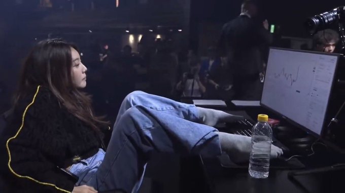 A side profile of a young woman with long dark hair, wearing a black sweater and blue jeans, reclining in a chair in a dimly lit event space with a laptop displaying charts and a water bottle nearby.