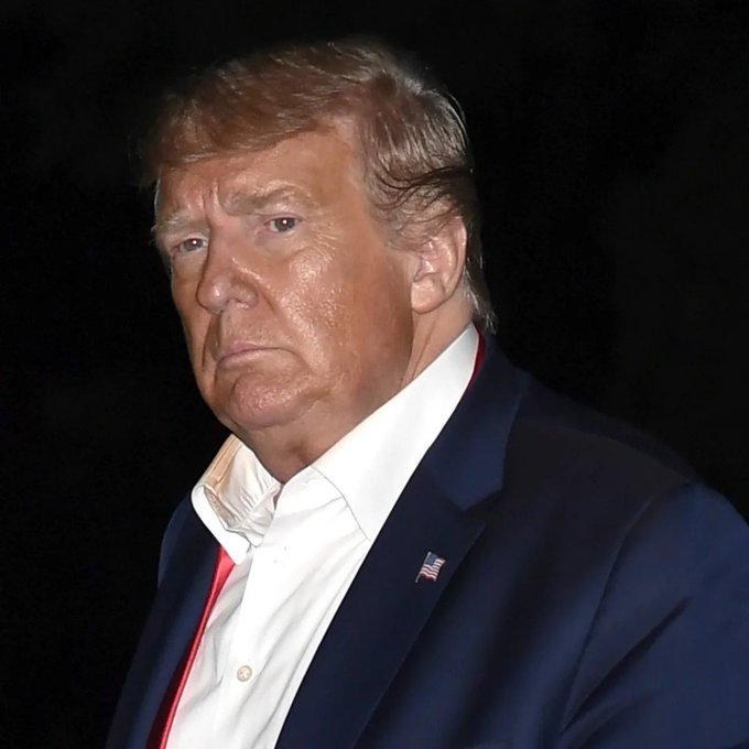 A close-up portrait of Donald Trump wearing a dark suit and white shirt with a US flag lapel pin against a black background.
