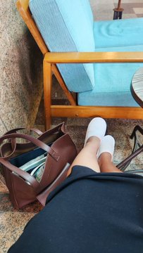 A first-person view looking down at white sneakers, a dark skirt, and a brown tote bag resting on the ground next to a wooden chair with blue cushions.