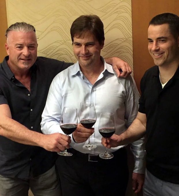 Three men stand closely together, each holding a glass of red wine, with the central figure in a light blue button-down shirt flanked by two men in dark shirts, capturing a moment of camaraderie that the post suggests represents an industry trio whose influence and shared memories remain largely unrecognized by many.